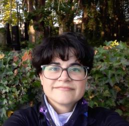 Photo of Celia rodrigues, a women wearing glasses with short hair standing in front of wall of ivy, and trees
