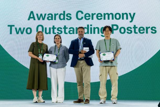Two poster prize winners stand on the stage with the conference hosts, holding their certificates. Text in the background reads "Awards ceremony: Two outstanding posters."