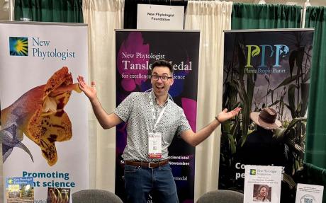 Bennett Young, looking excited, stands behind a table covered with journal articles, leaflets, pens, badges and other promotional material. The table cloth displays the logos of New Phytologist; Plants, People, Planet; and the New Phytologist Foundation.