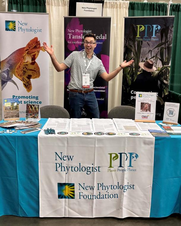 Bennett Young, looking excited, stands behind a table covered with journal articles, leaflets, pens, badges and other promotional material. The table cloth displays the logos of New Phytologist; Plants, People, Planet; and the New Phytologist Foundation.