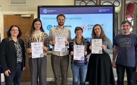Four poster prize winners stand with the certificates at in front of a display screen displaying sponsor logos at a conference.