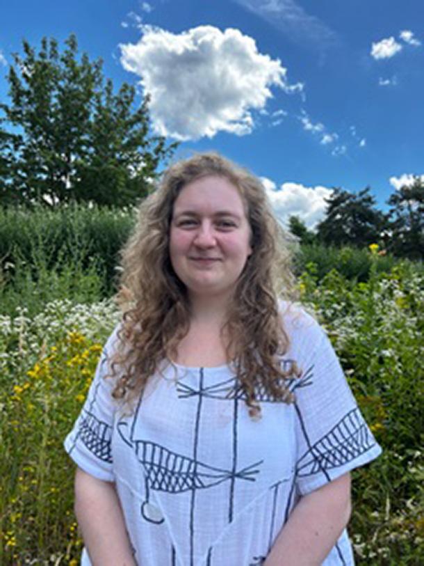 Photograph of Claire Scheid. In the background is a botanically diverse field boundary and a blue sky with scattered clouds.