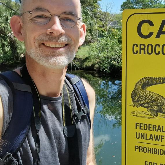 Jens-Christian Svenning stood smiling in front of a lake and bushes, next to a yellow warning sign that reads "caution crocodiles in area"