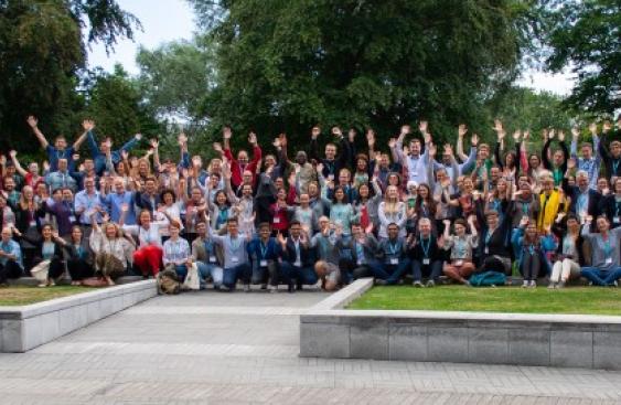 Group picture of the over 100 attendees at the Next Generation Scientists event at University College Dublin, Ireland in 2019. There are four rows of smiling people with their hands in the air in front of an area of trees.