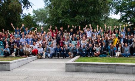 Group picture of the over 100 attendees at the Next Generation Scientists event at University College Dublin, Ireland in 2019. There are four rows of smiling people with their hands in the air in front of an area of trees.