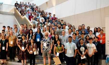 Group picture of the over 100 attendees of the Next Generation Scientists event at the National University of Singapore in 2023. The attendees are smiling and standing on a large staircase. A banner is to the right with the New Phytologist next generation scientists logo: three green circles, depicting a plant cell, two leaves and a globe and the New Phytologist Foundation logo: a blue and yellow sunbeam.
