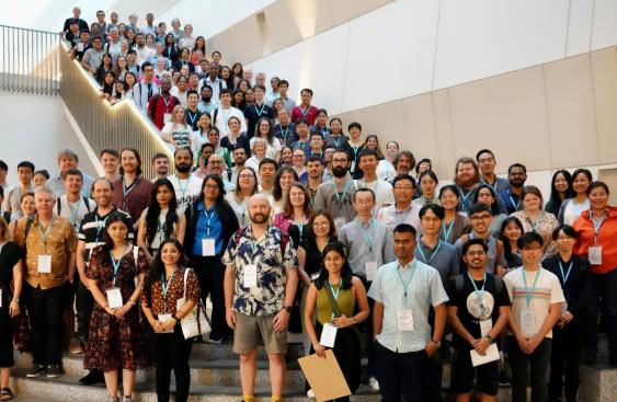 Group picture of the over 100 attendees of the Next Generation Scientists event at the National University of Singapore in 2023. The attendees are smiling and standing on a large staircase. A banner is to the right with the New Phytologist next generation scientists logo: three green circles, depicting a plant cell, two leaves and a globe and the New Phytologist Foundation logo: a blue and yellow sunbeam.