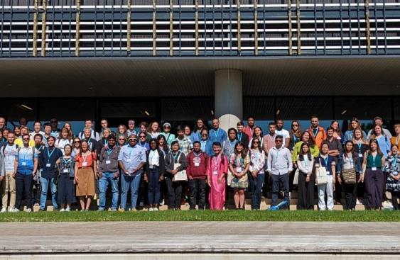 Group picture of the over 100 attendees of the Next Generation Scientist event at the University of Tartu in 2022. Three rows of attendees are stood on grass in front of a large building with three pillars. A banner is to the left with the New Phytologist next generation scientists logo: three green circles, depicting a plant cell, two leaves and a globe, and the New Phytologist Foundation logo: a blue and yellow sunbeam.