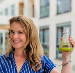 Photo of Joanna  Kacprzyk. A women with dark blond hair, smilling and holding a beaker of something green.  She is wearing a blue dress with white stripes