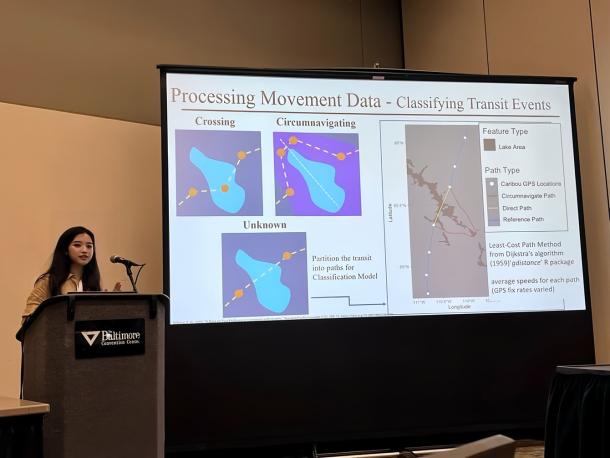 Qianru Liao stands behind a lectern as she delivers a scientific presentation. To her left is a presentation screen displaying a slide including diagrams and maps.