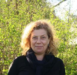 Maria E. Eriksson wearing a black coat, stood in front of bushes