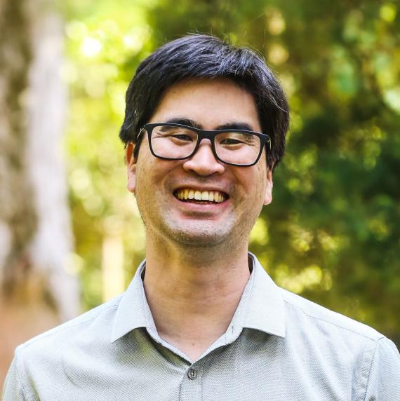 Carlos	Hotta wearing glasses and a grey shirt stood outside, smiling