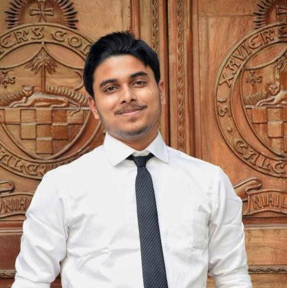Kankan Datta wearing a shirt and tie, stood infront of a wooden background with crests