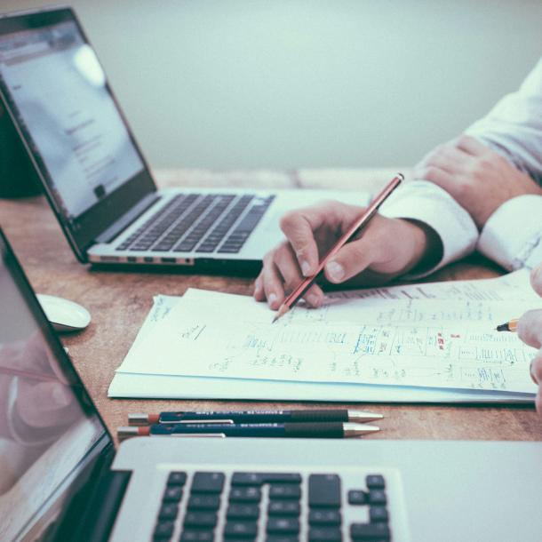 Two people work on a document in between two laptops, using pens and pencils. Only their hands are visible.