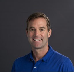 Josh Gendron wearing a blue polo smiling, in front of a dark grey background
