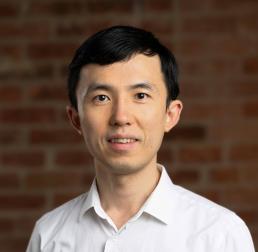Kai Zhu, wearing a white shirt in front of a red brick wall
