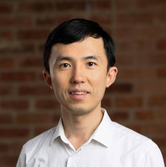 Kai Zhu, wearing a white shirt in front of a red brick wall